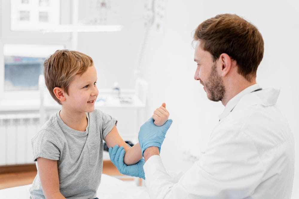 A young boy sits on a treatment bed, smiling as a healthcare professional in a white coat gently examines his arm. They are in a bright, clean medical room, creating a positive environment.