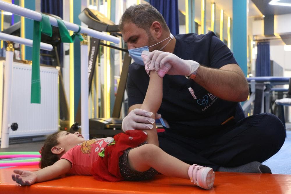 A healthcare professional in a blue shirt kneels beside a young girl on an exercise mat. He is gently holding her leg while stretching it, focusing on her movement. The background includes rehabilitation equipment, enhancing the clinical setting.