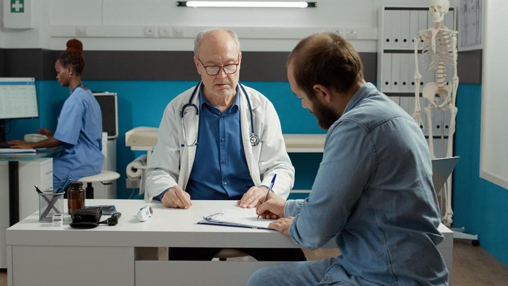 A doctor with gray hair and glasses is seated at a desk, reviewing a file while a male patient is taking notes. A nurse in the background is working on a computer. The office is well-equipped, featuring medical tools and anatomical charts on the walls.