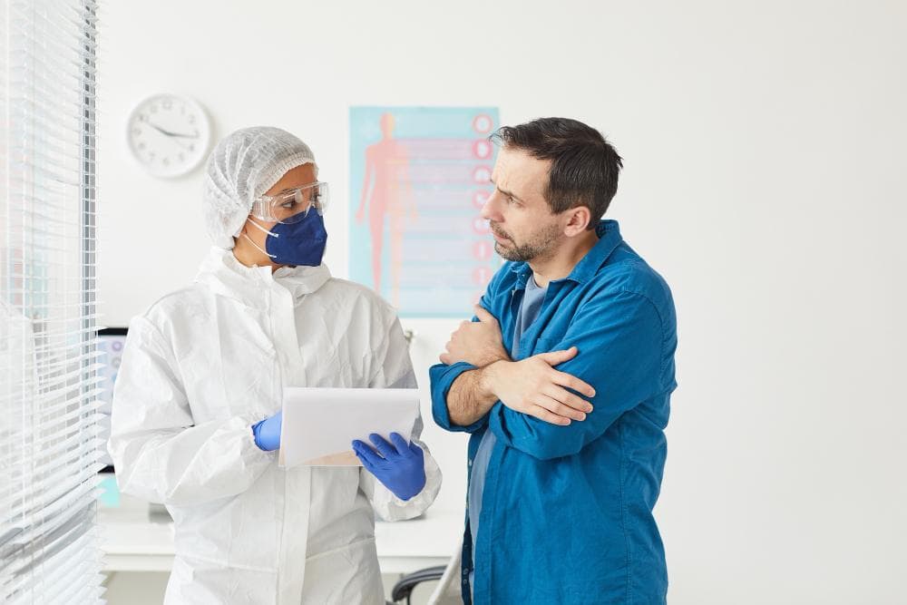 A healthcare worker in full protective gear, including a mask and face shield, is discussing medical information with a male patient. The doctor is holding a clipboard with notes while the patient, with arms crossed, looks contemplative.