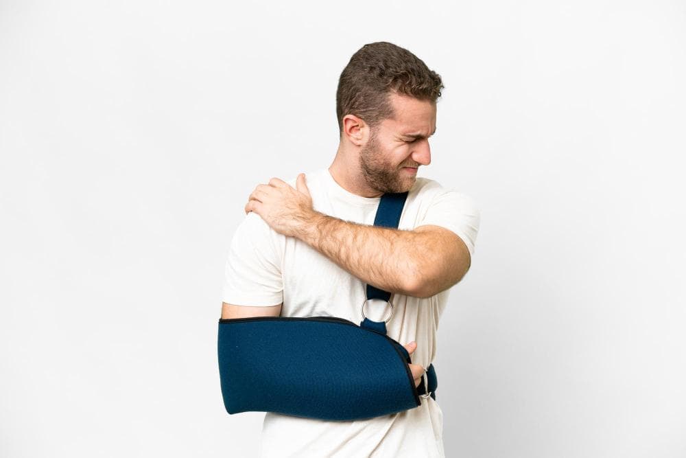 A young man with short brown hair is seen wincing in pain while holding his shoulder with one hand. He is wearing a white t-shirt and has a blue arm sling that supports his injured arm. The background is plain white.